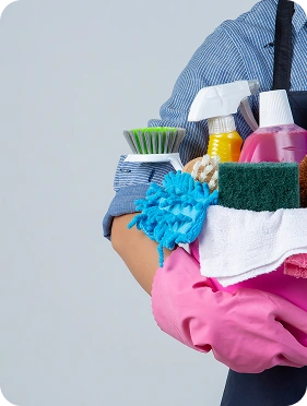 Osoba ubrana w niebieską koszulę i fartuch trzyma różowe wiadro ze środkami czyszczącymi, takimi jak spraye, szczotki i ściereczki. Ma na sobie różowe gumowe rękawiczki i jest gotowa do sprzątania.